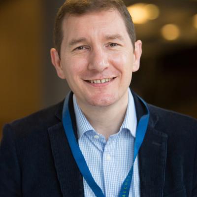 A photo of Cormac with a light blue shirt and a dark jacket with a blue lanyard 